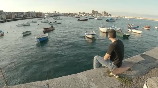 穿着蓝色牛仔裤和黑色短袖T恤的小伙子日落时坐在码头上沉思 — 图库视频影像