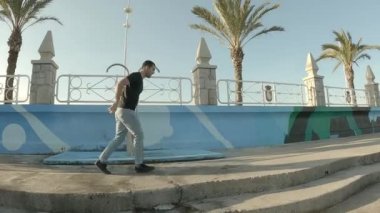 Kotlar ve siyah kısa kollu tişörtler giyen genç bir çocuk güneşli bir öğleden sonra deniz kenarında paten kayıyor.