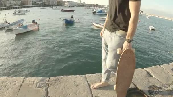 若い男がジーンズと黒の半袖Tシャツを着て裸足で歩くと 日没時に桟橋に沿ってスケートボード — ストック動画
