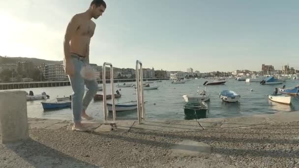 若いです男に身を包んだジーンズと無気力な散歩裸足でとともにザ 桟橋に沿ってスケートボードで日没 — ストック動画