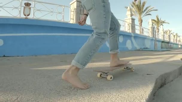 晴れた日の午後に海辺の遊歩道に沿って裸足でスケートTシャツのないジーンズを着た若い男の子 — ストック動画