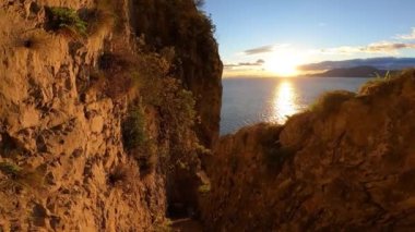 Gün doğumunda denize bakan kayalık bir uçurumdan aşağı iniyor. Çevrede bitkiler