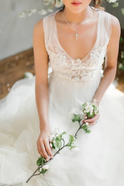 A girl in a white wedding dress holds in her hands a flowering branch of an apple tree. Hands of the bride close-up. On the neck of the girl a chain with a cross. Scarlet lips. Tulle wedding dress