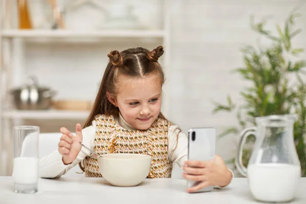 Маленька дівчинка снідає і дивиться на смартфон — стокове фото
