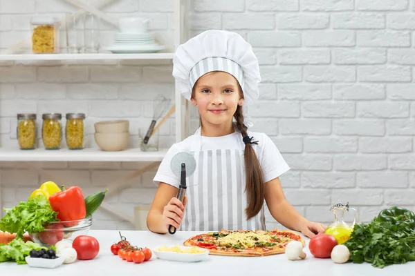 Nettes kleines Mädchen mit Kochmütze und einer Schürze, die Pizza schneidet — Stockfoto