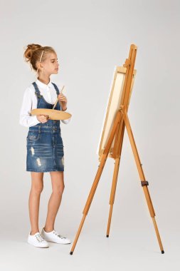 Güzel küçük bir kızın portresi ahşap bir palet ve stüdyo arka planında fırça taşıyor.