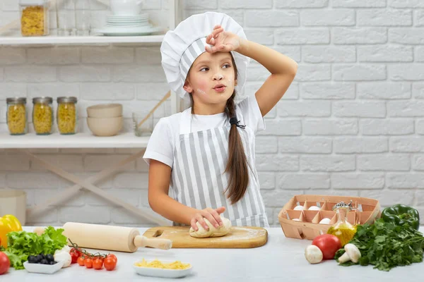 Kislány sapkában és kötény gyúrja a tésztát. — Stock Fotó