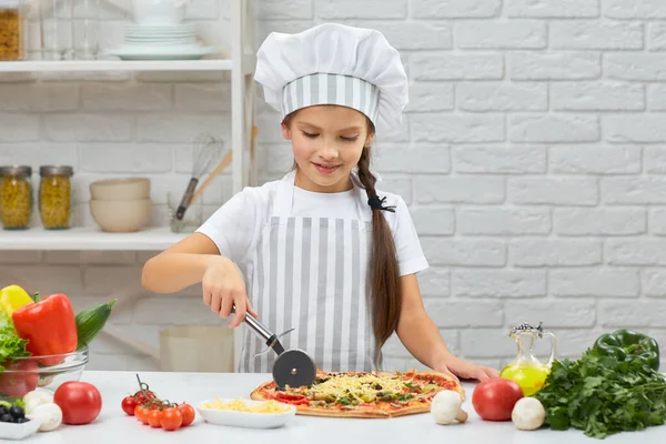 Nettes kleines Mädchen mit Kochmütze und einer Schürze, die Pizza schneidet — Stockfoto
