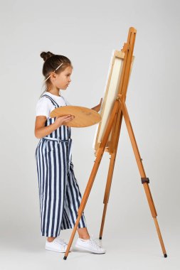 Güzel küçük bir kızın portresi ahşap bir palet ve stüdyo arka planında fırça taşıyor.