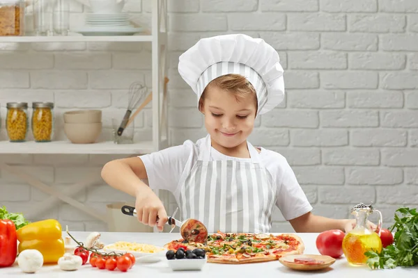 Egy kisfiú séf kalapban és egy kötény főzés pizza — Stock Fotó