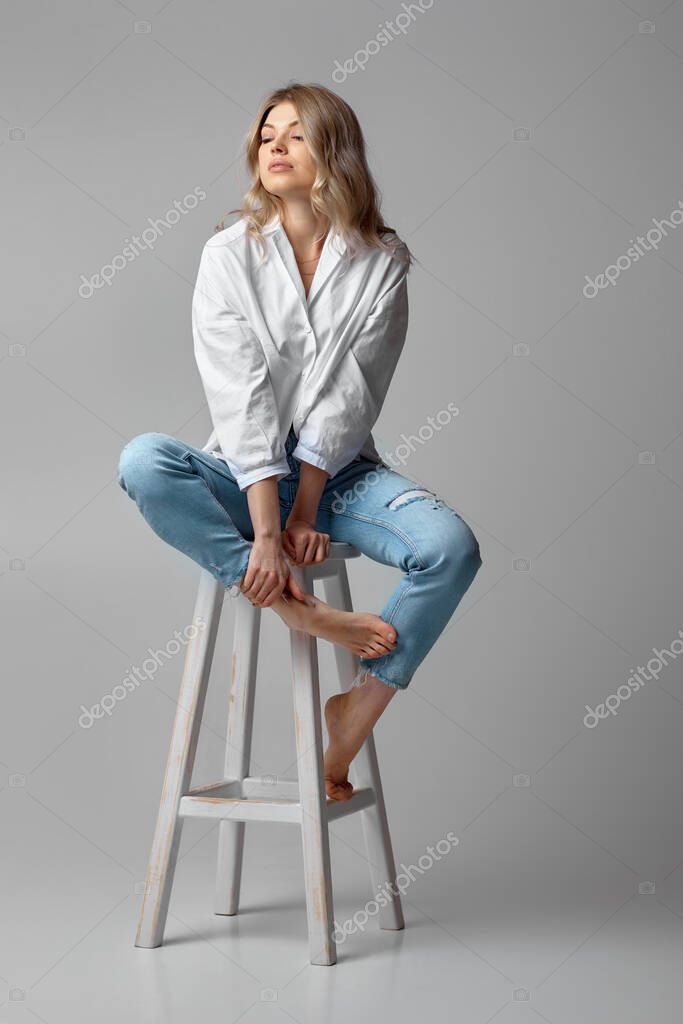 Beautiful Blonde Woman With Curly Hair In Jeans And A White Shirt Sitting On Chair On Studio Background Larastock Beautiful Blonde Woman With Curly Hair In Jeans And A White Shirt Sitting On Chair On Studio Background Larastock