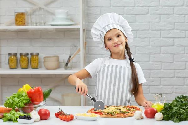 Милая маленькая девочка в шляпе шеф-повара и фартуке, режет пиццу. — стоковое фото