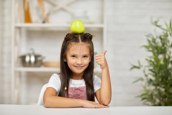 Маленька дівчинка сидить за столом з яблуком на голові — стокове фото