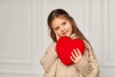 Kırmızı kalbi beyaz bir duvara dayayan küçük tatlı bir kız çocuğu..