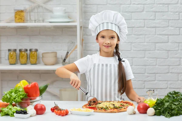 Nettes kleines Mädchen mit Kochmütze und einer Schürze, die Pizza schneidet — Stockfoto