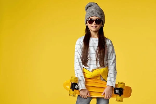 Дитяча дівчина в капелюсі і сонцезахисних окулярах з жовтим скейтбордом — стокове фото