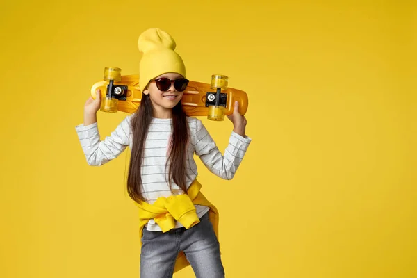 Дитяча дівчина в капелюсі і сонцезахисних окулярах з жовтим скейтбордом — стокове фото