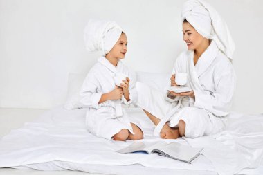 Anne ve kızı beyaz bornozlu çay içiyorlar.