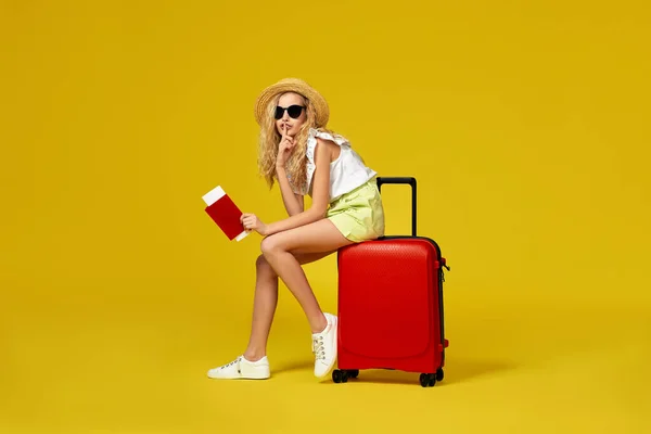 Дівчина в капелюсі з паспортом і квитком, що сидить на червоній валізі — стокове фото