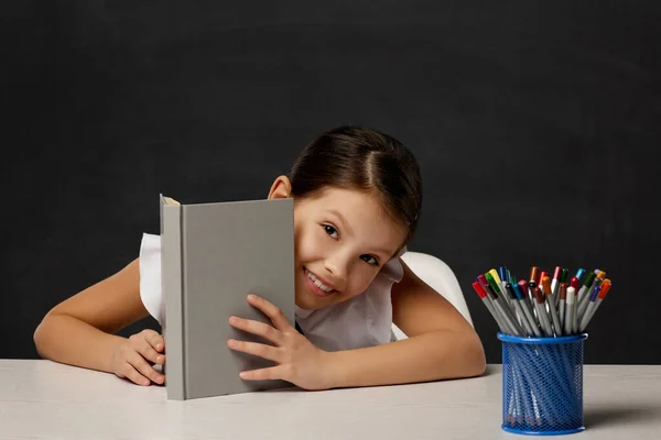 Klein schoolmeisje kijkt uit van achter het boek — Stockfoto