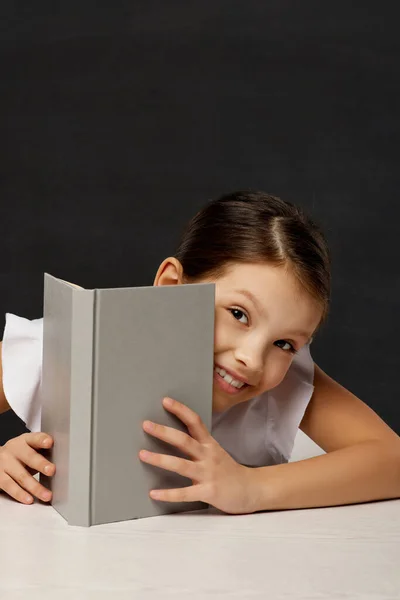 Klein schoolmeisje kijkt uit van achter het boek — Stockfoto