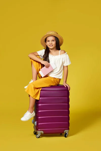Дитяча дівчина в капелюсі з паспортом і квитком, що сидить на валізі — стокове фото