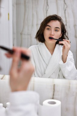 Beyaz bornozlu kadın aynanın önünde dişlerini fırçalıyor.