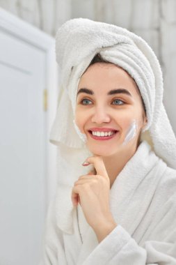 Beyaz bornozlu kadın yüz kremi sürüyor.