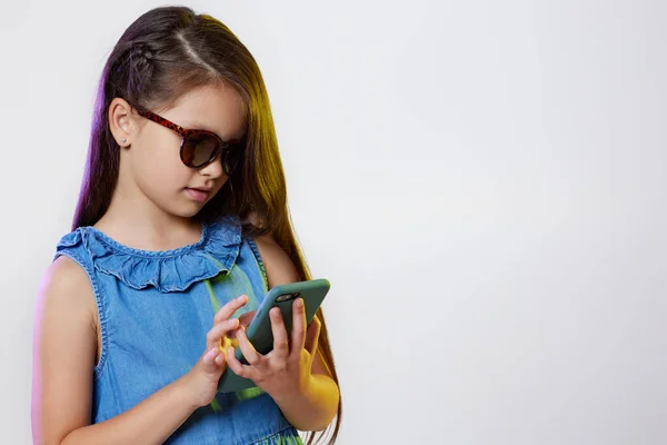 Gyermek lány napszemüveg segítségével okostelefon fehér háttér — Stock Fotó