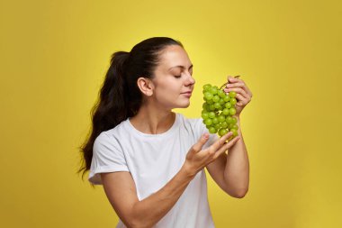 Çekici beyaz kadın portresi sarı stüdyo arka planında izole edilmiş üzümler tutuyor.