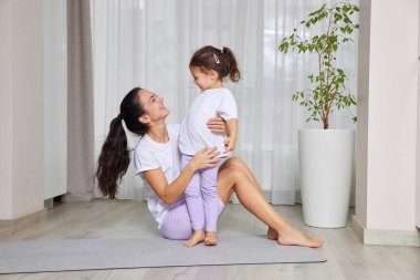 Anne ve kızı egzersiz yaparken ve yoga minderinde oynarken gülümsüyor.