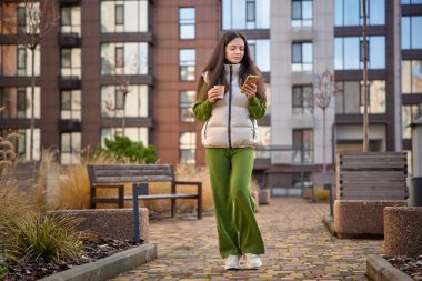 Genç bir kadın dışarıda yürüyor, elinde bir kahve fincanı ile akıllı telefonuna bakıyor.
