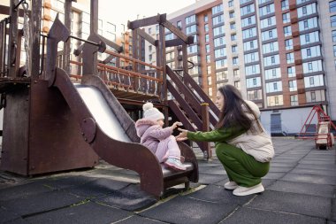 Anne, şehir parkında konut binaları olan bir çocuğa kaydırağında yardım ediyor.
