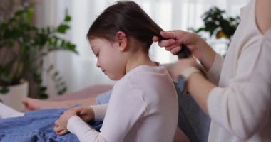 Genç bir kadın, sevimli kızlarını uzun siyah saçlarıyla kanepede at kuyruğu şeklinde şekillendiriyor.