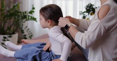 Genç bir kadın, sevimli kızlarını uzun siyah saçlarıyla kanepede at kuyruğu şeklinde şekillendiriyor.