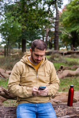 Genç ve çekici bir adamın ağaçta oturup telefonuna bakarken doğa gezisine ara verdiği bir fotoğraf. Ormandaki ağaçlarımın etrafında.