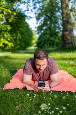 Genç ve çekici bir adamın parkta bir havlunun üzerinde uzanmış akıllı telefonunu kontrol ederken dışarıda geçirdiği yaz gününün keyfini çıkarırken fotoğrafı.. 