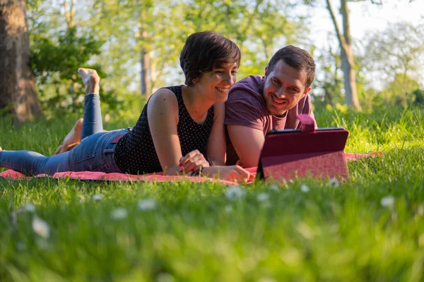 Photo of a young and attractive couple lying on a towel in the park watching a video on an electronic device and smiling at each other. Great summer day, relaxed attitude.