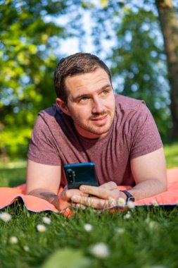 Genç ve çekici bir adamın parkta bir havlunun üzerinde uzanmış akıllı telefonunu kontrol ederken dışarıda geçirdiği yaz gününün keyfini çıkarırken fotoğrafı.. 