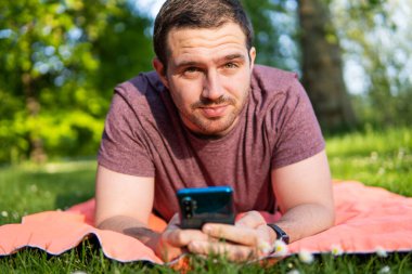 Genç ve çekici bir adamın parkta bir havlunun üzerinde uzanmış akıllı telefonunu kontrol ederken dışarıda geçirdiği yaz gününün keyfini çıkarırken fotoğrafı.. 