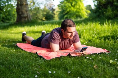 Genç ve çekici bir adamın parkta bir havlunun üzerinde uzanmış akıllı telefonunu kontrol ederken dışarıda geçirdiği yaz gününün keyfini çıkarırken fotoğrafı.. 