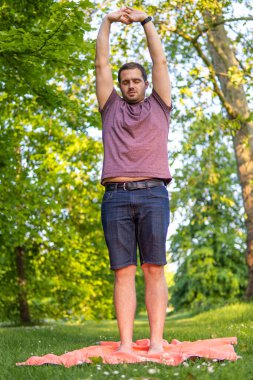 Parkta kolları gökyüzüne kadar uzanan ve gözleri kapalı genç ve çekici bir adamın fotoğrafı. Bir yaz günü doğada tek başına. Yoga pozu