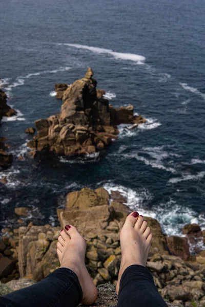 Photo of feet of a woman with red nails sitting on a rock in front of a beautiful landscape next to the sea. Amazing natural beach in UK. Lands End. Travelling in a sunny day
