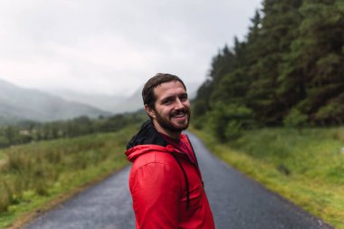 İskoçya, İskoçya 'da bulutlu bir günde bir ormanla çevrili boş bir yolda yürüyen kırmızı yağmurluklu bir adam.