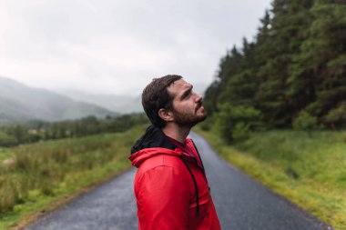 İskoçya, İskoçya 'da bulutlu bir günde bir ormanla çevrili boş bir yolda yürüyen kırmızı yağmurluklu bir adam.