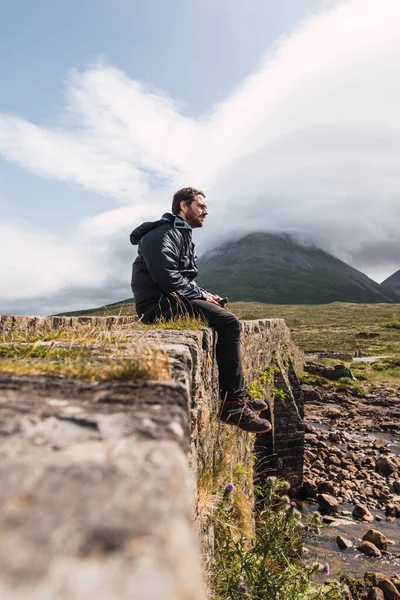 Young man sitting on the rock bridge while enjoying Scotland hill view at the Highlands, in Scotland. He is wearing hiking clothes. Sitting on an old vintage bridge in Sligachan, Isle of Skye