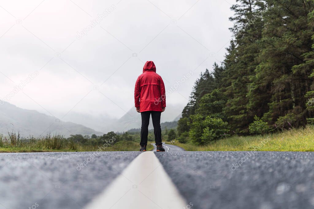 Traveler walking into the horizon on the lonely road in Scotland enjoying the scenery. He is wearing a red raincoat and surrounded by green trees and nature. Enjoying freedom