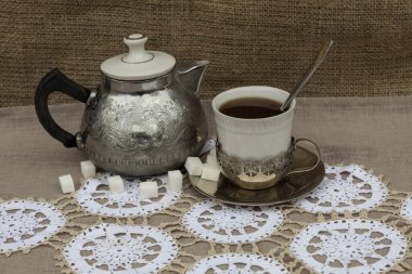 Beyaz bir peçetede, bardak tutacağında bir fincan çay ve metal bir çaydanlık var. Yakınlarda küp şekerler var.