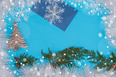 Noel süsleri, kozalaklı dallar, kar taneleri, bir zarf ve parlak bir Noel ağacı mavi arka planda çerçevelenmiştir..