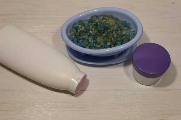 There is a soap dish in the form of a bathtub filled with blue salt on a white background. Nearby is a jar of skin cream and a bottle of cosmetic product.